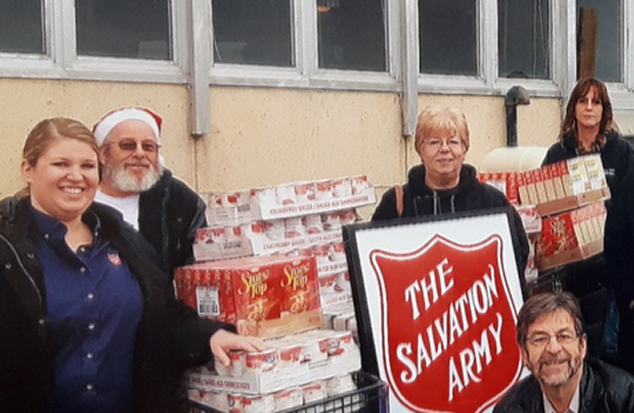 Group of the Salvation Army staff and volunteers standing together with donated food boxes and a Salvation Army shield sign outside a building.